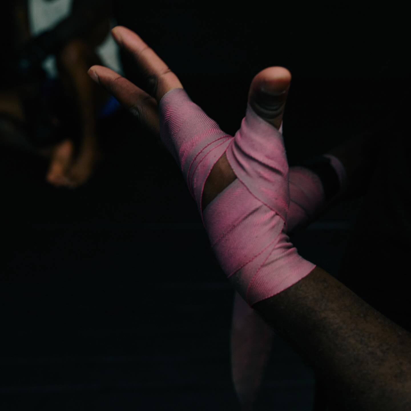 Closeup of a dark-skinned person's hand and arm wrapped in pink bandage; arm is raised in a dark indoor environment.