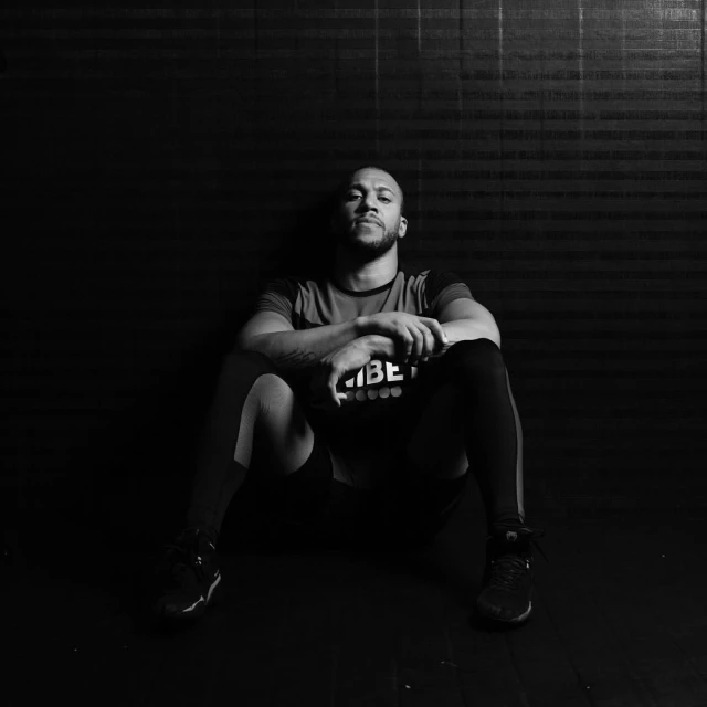 Monochrome portrait of a man with short hair seated against a textured wall, wearing athletic wear and sneakers in a studio setting.