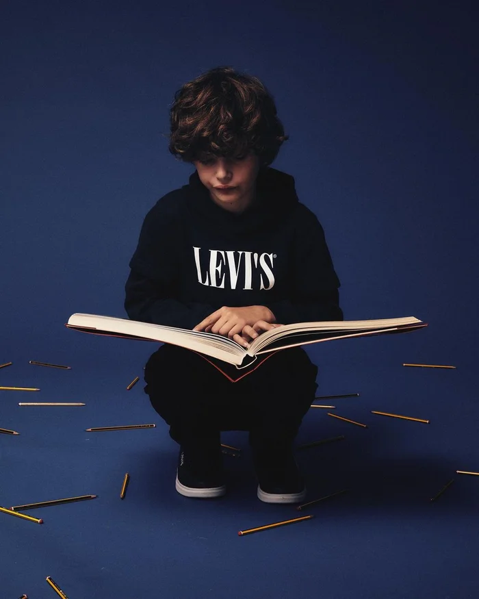 Young boy with curly brown hair crouching while reading a large open book against a solid dark blue studio backdrop with scattered pencils.