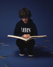 Young boy with curly brown hair crouching while reading a large open book against a solid dark blue studio backdrop with scattered pencils.
