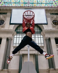 A woman in a red jacket and dark pants hanging from a basketball hoop inside an ornate building with glass windows.