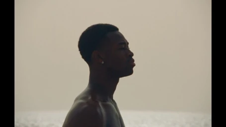 Profile portrait of a shirtless Black man outdoors against an overcast sky and a calm body of water.