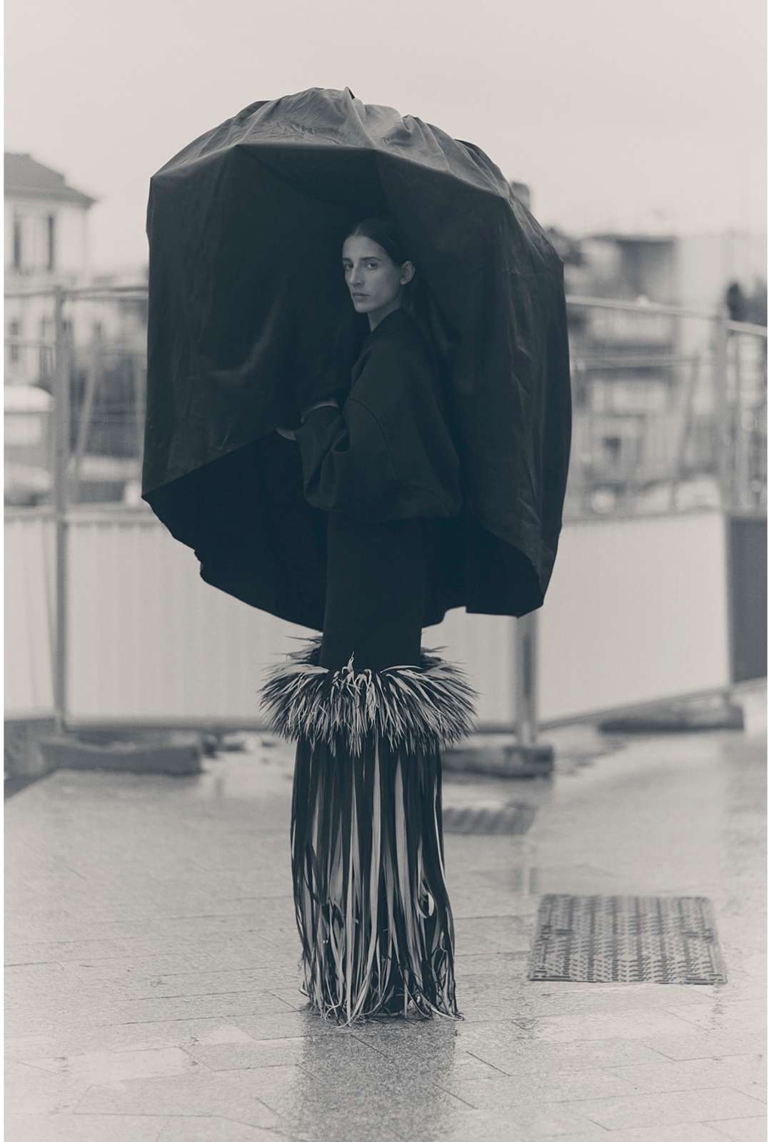 A woman stands under an umbrella wearing a fringed skirt on a wet city street, captured in a black-and-white medium shot with overcast lighting.