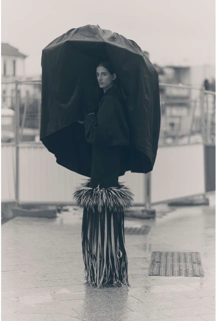 A woman stands under an umbrella wearing a fringed skirt on a wet city street, captured in a black-and-white medium shot with overcast lighting.