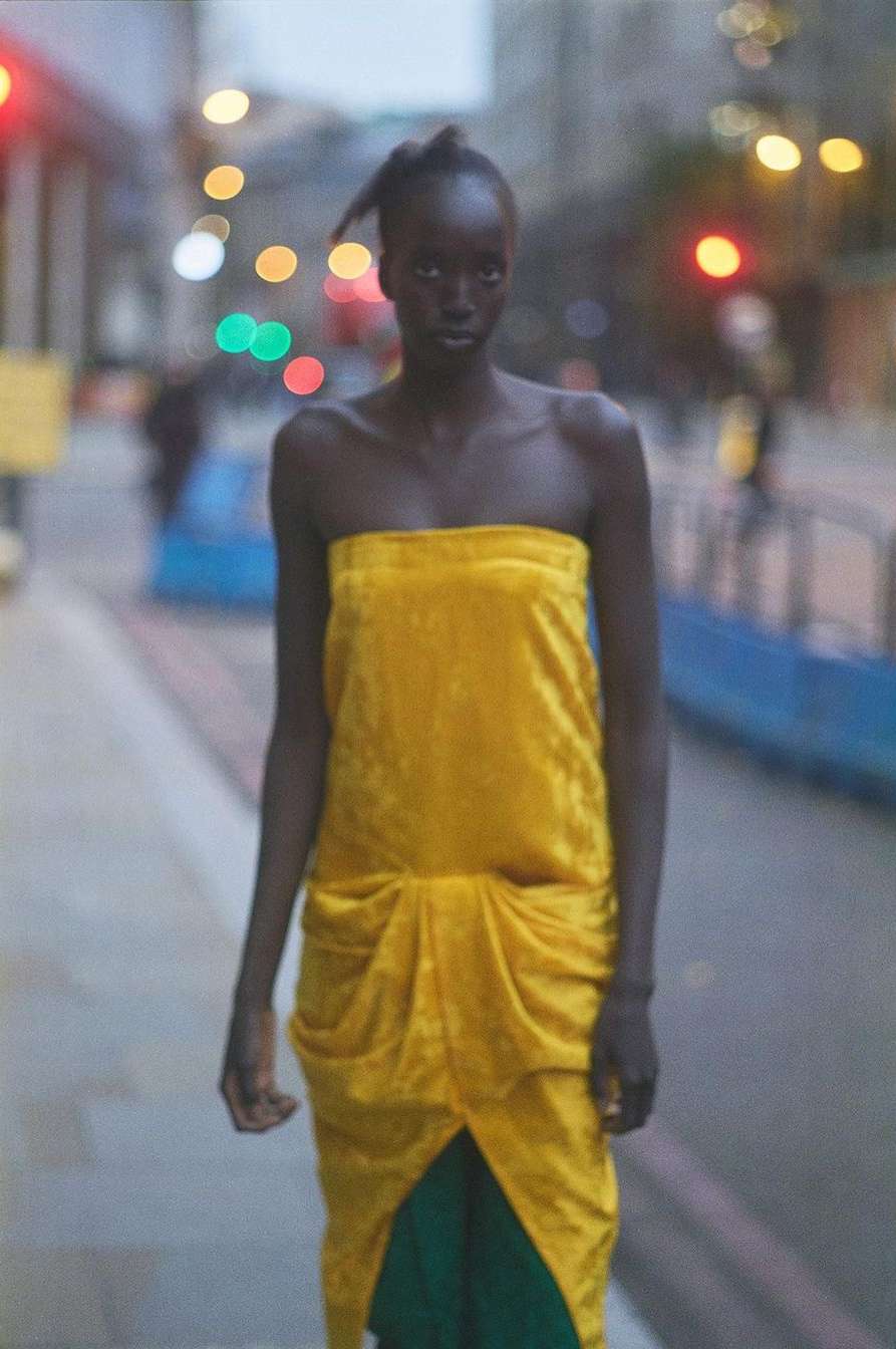 Woman with dark skin and a yellow dress stands on a city street in soft lighting, posing for fashion or editorial photography.