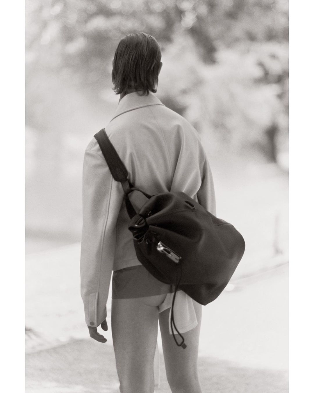 Monochrome fashion shot from behind of a person with a black drawstring bag, wearing a jacket and shorts, in a blurred nature setting.