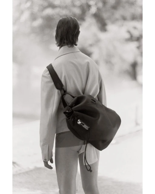 Monochrome fashion shot from behind of a person with a black drawstring bag, wearing a jacket and shorts, in a blurred nature setting.