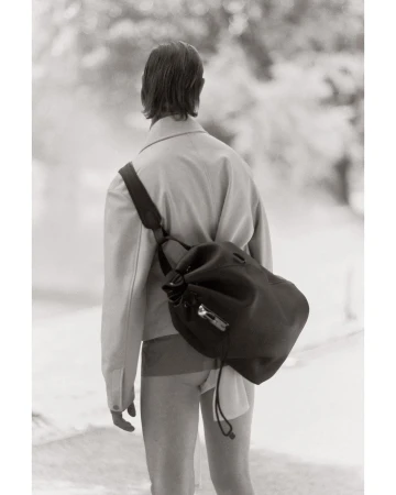 Monochrome fashion shot from behind of a person with a black drawstring bag, wearing a jacket and shorts, in a blurred nature setting.