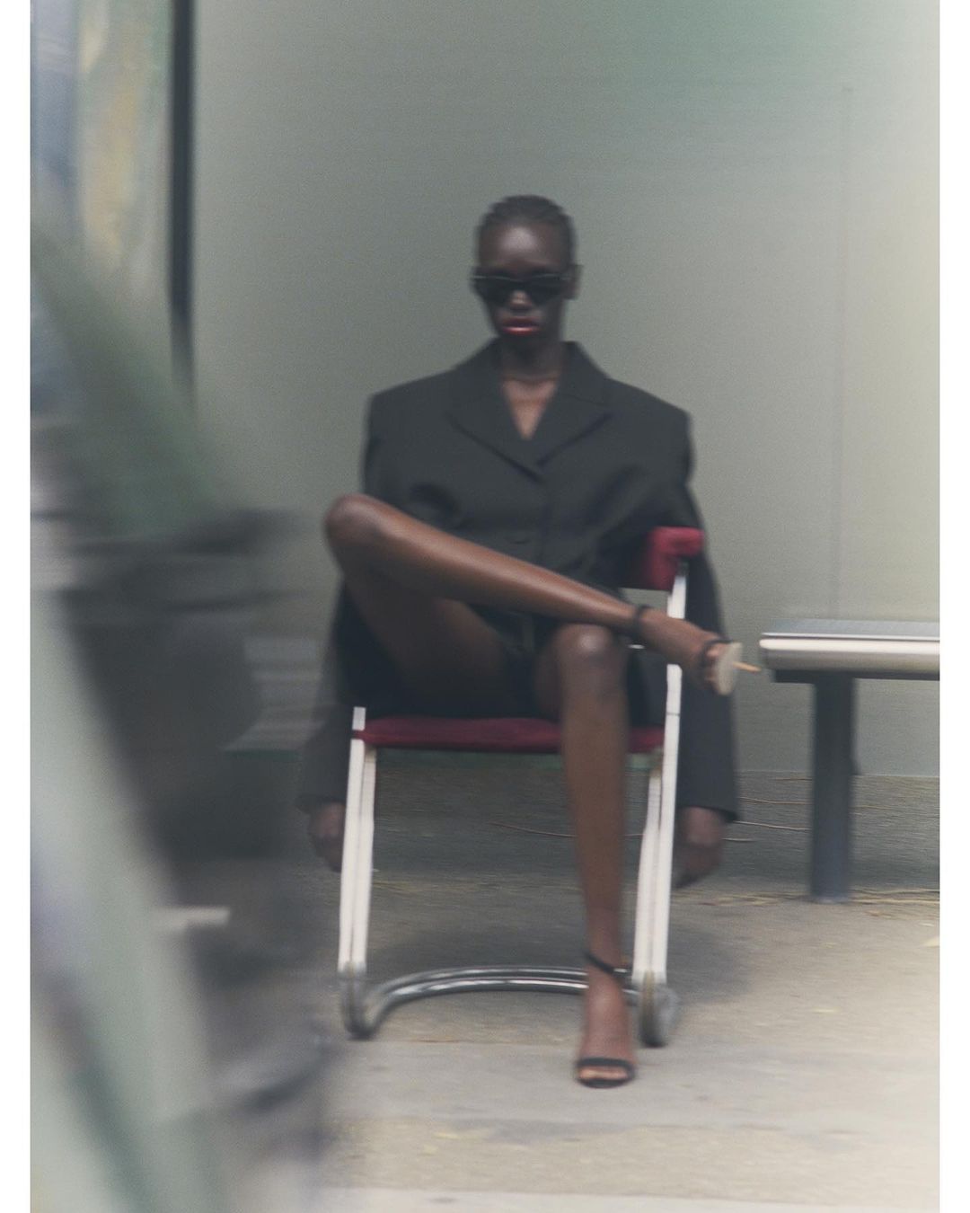 Elegant Black woman poses seated in a suit and heels, confidently crossing her legs, with blurry background, wearing dark sunglasses.
