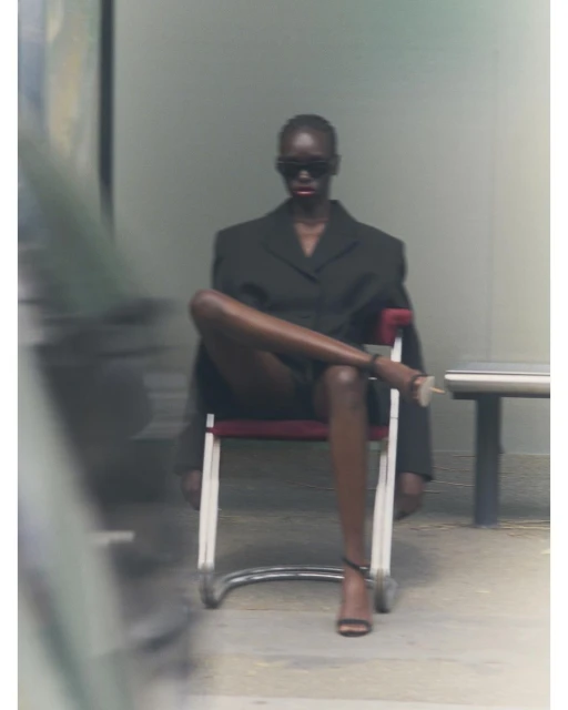 Elegant Black woman poses seated in a suit and heels, confidently crossing her legs, with blurry background, wearing dark sunglasses.