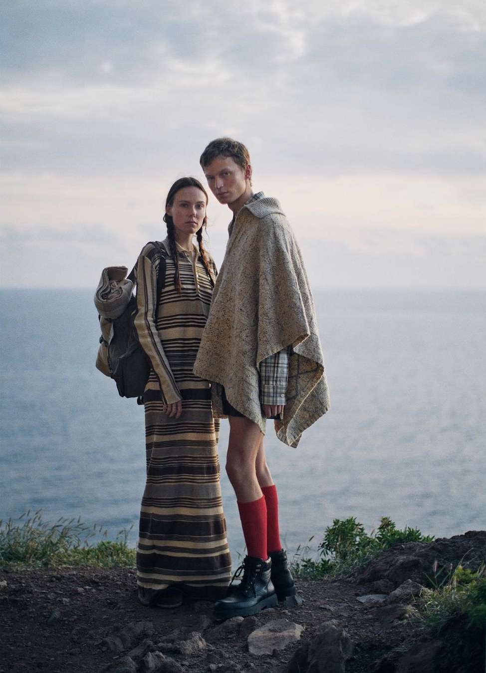 Two models standing on a rocky coast near the ocean, wearing a striped dress and a beige poncho with red socks.