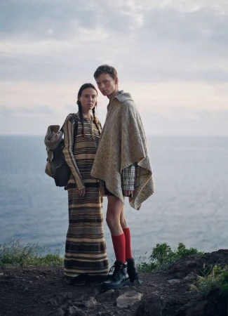 Two models standing on a rocky coast near the ocean, wearing a striped dress and a beige poncho with red socks.