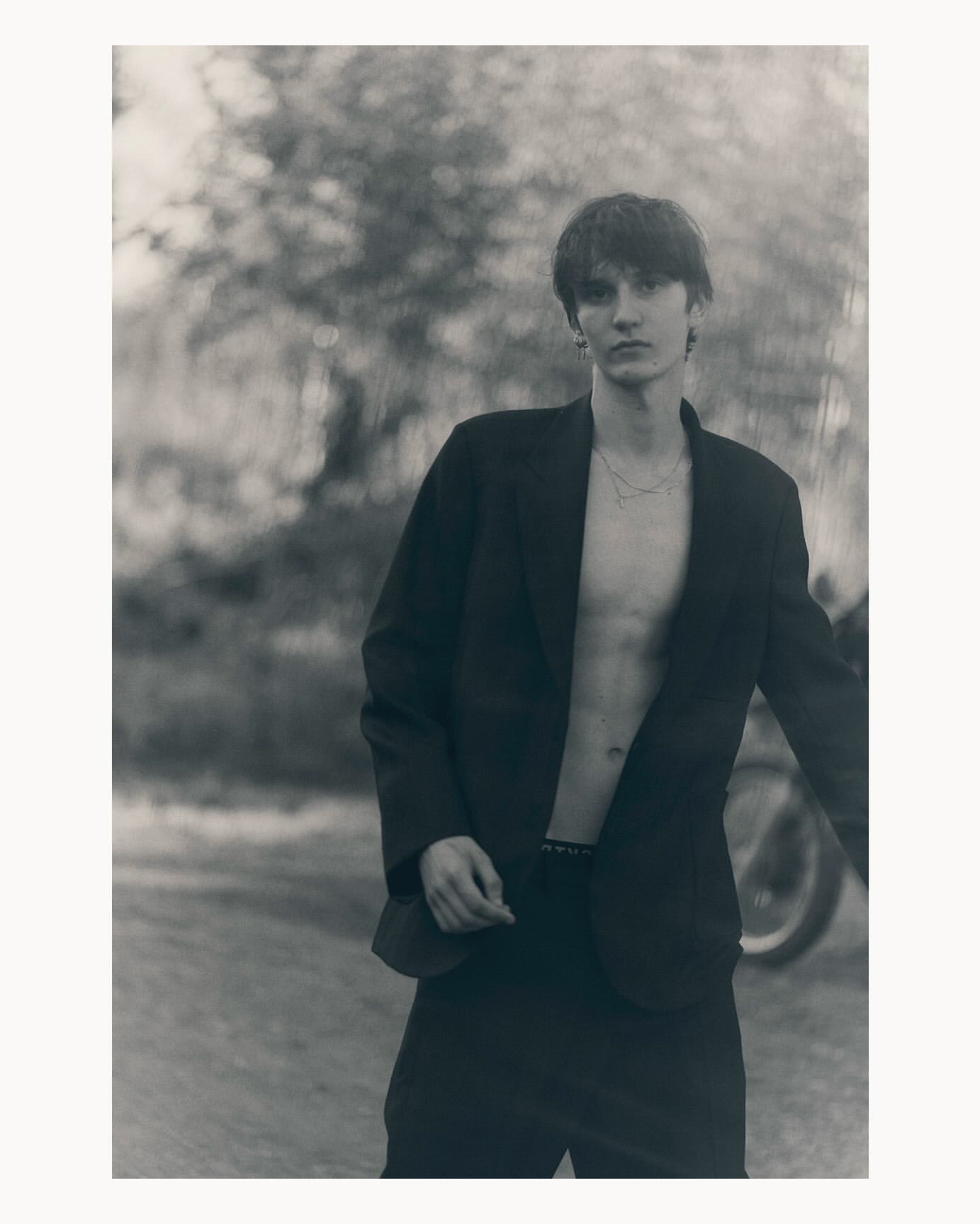 Young man in blazer stands outdoors against a blurred foliage backdrop, creating a striking black-and-white fashion portrait.