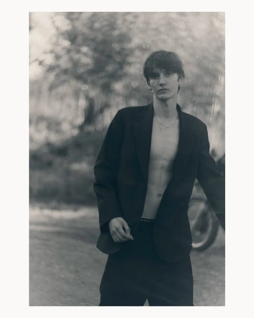 Young man in blazer stands outdoors against a blurred foliage backdrop, creating a striking black-and-white fashion portrait.