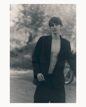 Young man in blazer stands outdoors against a blurred foliage backdrop, creating a striking black-and-white fashion portrait.