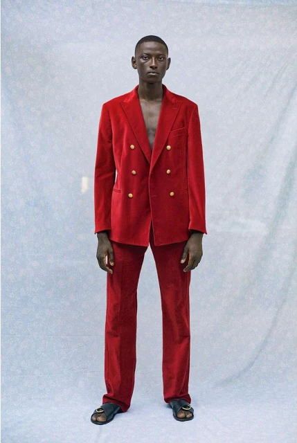 A slender, dark-skinned man poses in a vibrant red, double-breasted velvet suit with gold buttons and black leather sandals against a textured light blue backdrop.