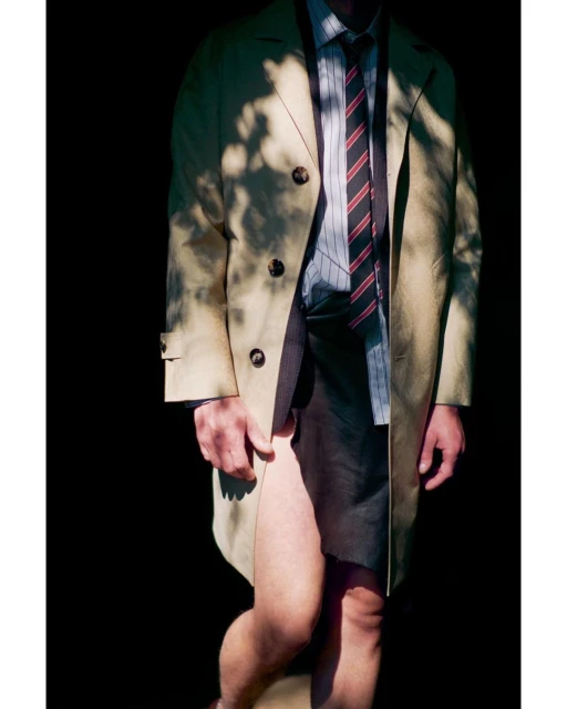 A person in a trench coat and striped tie poses in shadows, creating a strong contrast between light and dark.