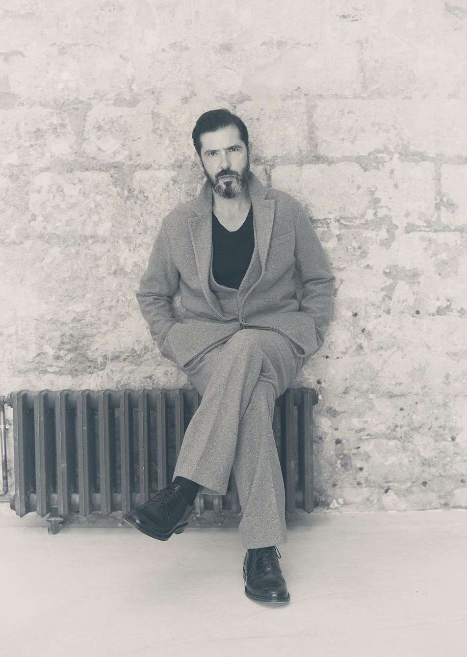 Monochrome portrait of a bearded adult male in a suit, seated against a textured brick wall, with a vintage radiator in the scene.