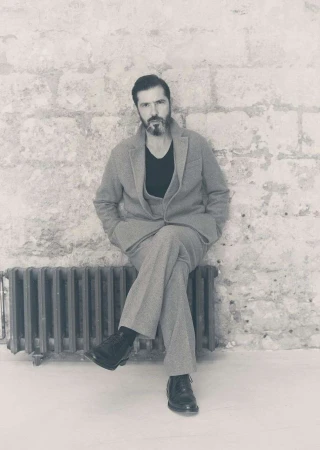 Monochrome portrait of a bearded adult male in a suit, seated against a textured brick wall, with a vintage radiator in the scene.