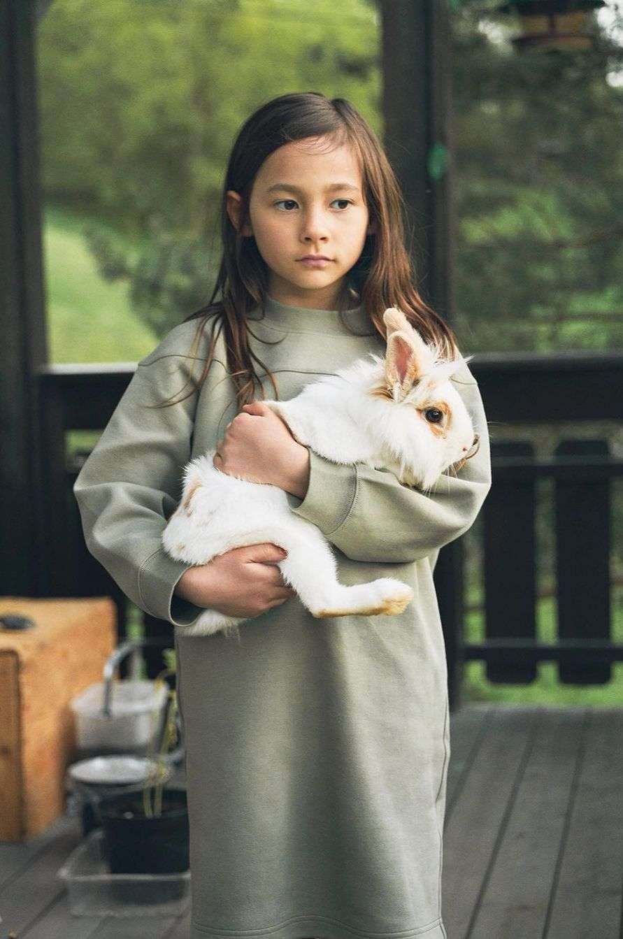 A young girl in a green dress stands outside, cradling a fluffy white rabbit in her arms, with a serene expression on her face.
