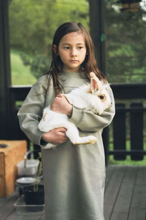 A young girl in a green dress stands outside, cradling a fluffy white rabbit in her arms, with a serene expression on her face.