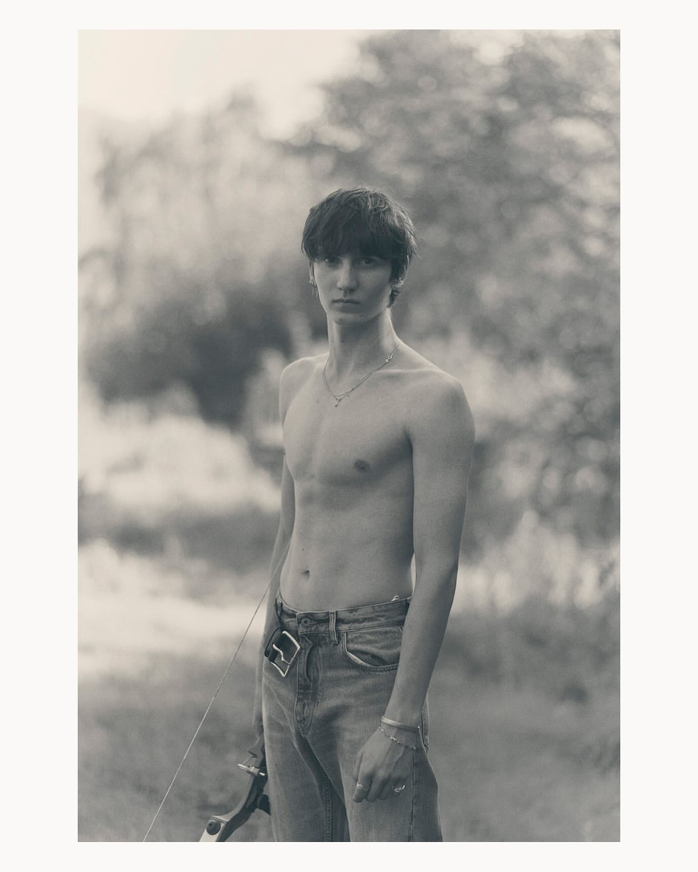 Monochrome portrait of a shirtless young man with an archery bow standing outdoors in front of blurred trees and natural lighting.