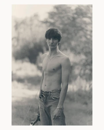 Monochrome portrait of a shirtless young man with an archery bow standing outdoors in front of blurred trees and natural lighting.