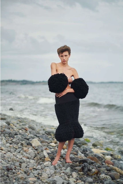 Woman poses in high-fashion skirt and arm cuffs on rocky beach, featuring neutral tones and overcast skies, with a portrait orientation.