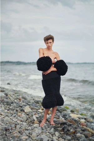Woman poses in high-fashion skirt and arm cuffs on rocky beach, featuring neutral tones and overcast skies, with a portrait orientation.