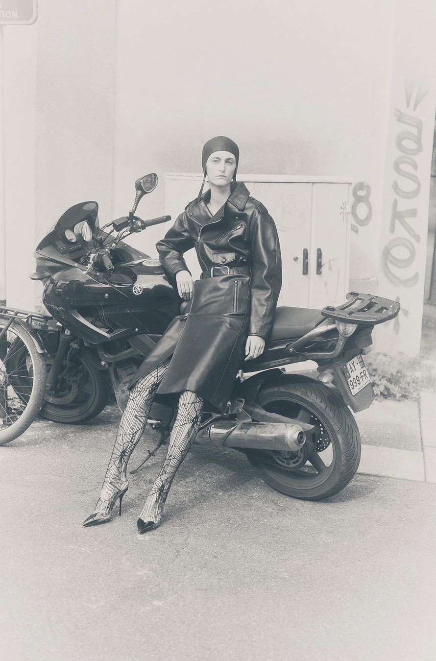 A monochromatic fashion shot of a woman in a leather skirt suit and printed tights sitting on a black motorcycle outdoors against a building.