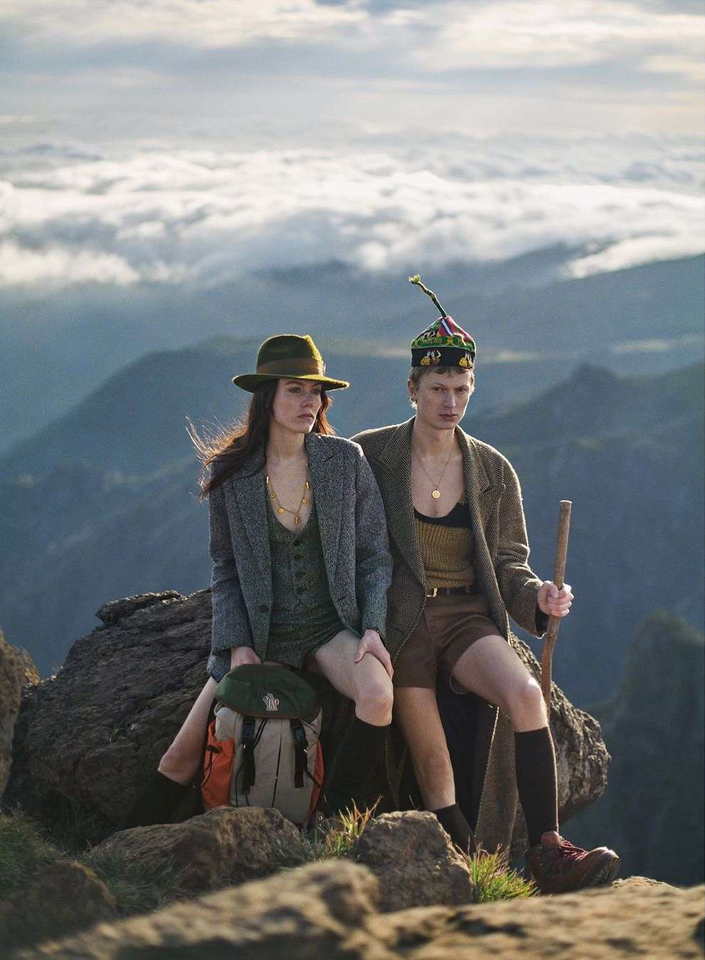 Two people in outdoor fashion shoot on rocky mountain with scenic cloudscape, wearing hats, jackets, and hiking boots.