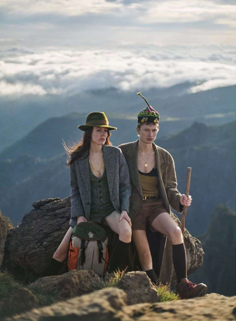 Two people in outdoor fashion shoot on rocky mountain with scenic cloudscape, wearing hats, jackets, and hiking boots.