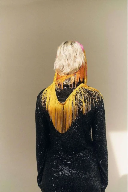 Back view of a female with blonde hair wearing a black sequined dress adorned with a striking gold fringe necklace, in a neutral setting.
