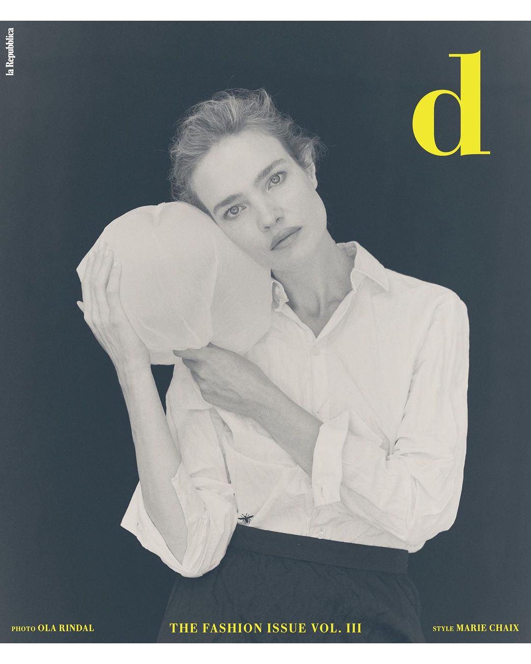 Fashion magazine cover featuring a fair-skinned adult female model in a white button-down shirt posing with a white object on a grey background.