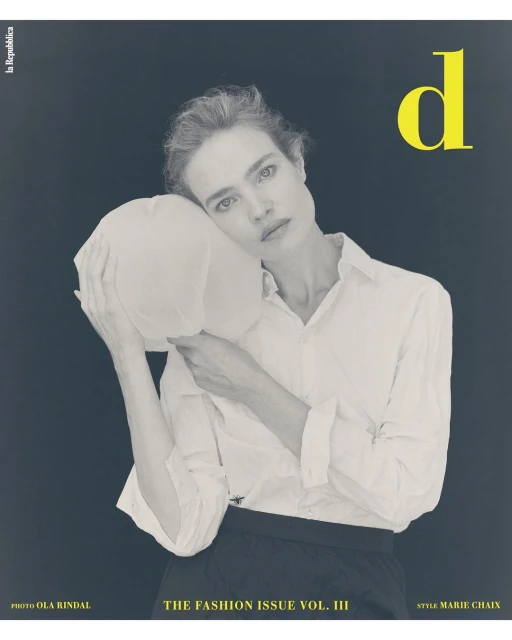 Fashion magazine cover featuring a fair-skinned adult female model in a white button-down shirt posing with a white object on a grey background.