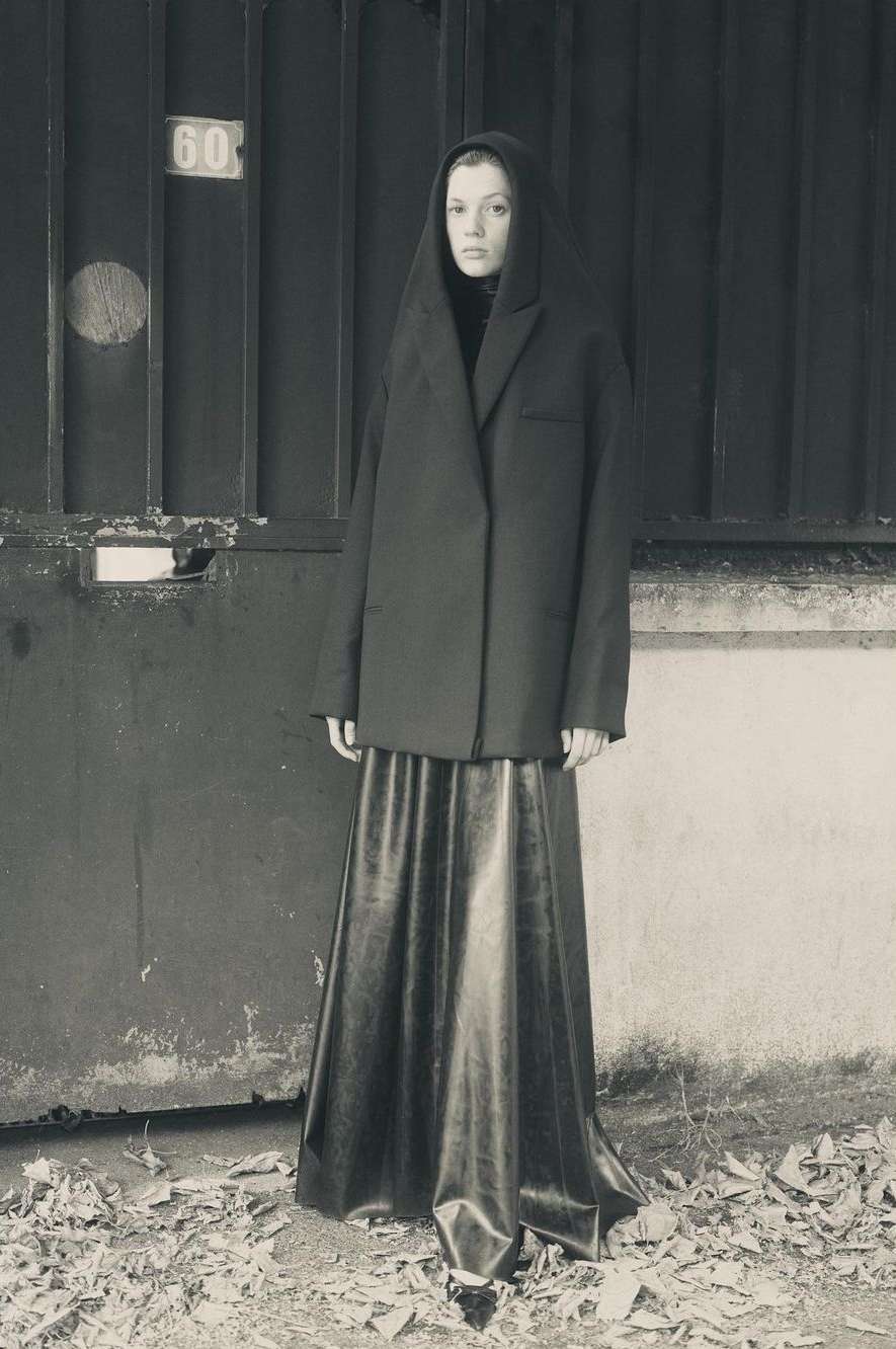 A woman stands in front of a metal gate, wearing a long, dark skirt and an oversized blazer with a hood up in a desaturated scene.