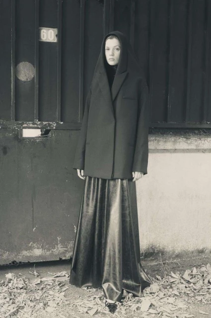 A woman stands in front of a metal gate, wearing a long, dark skirt and an oversized blazer with a hood up in a desaturated scene.