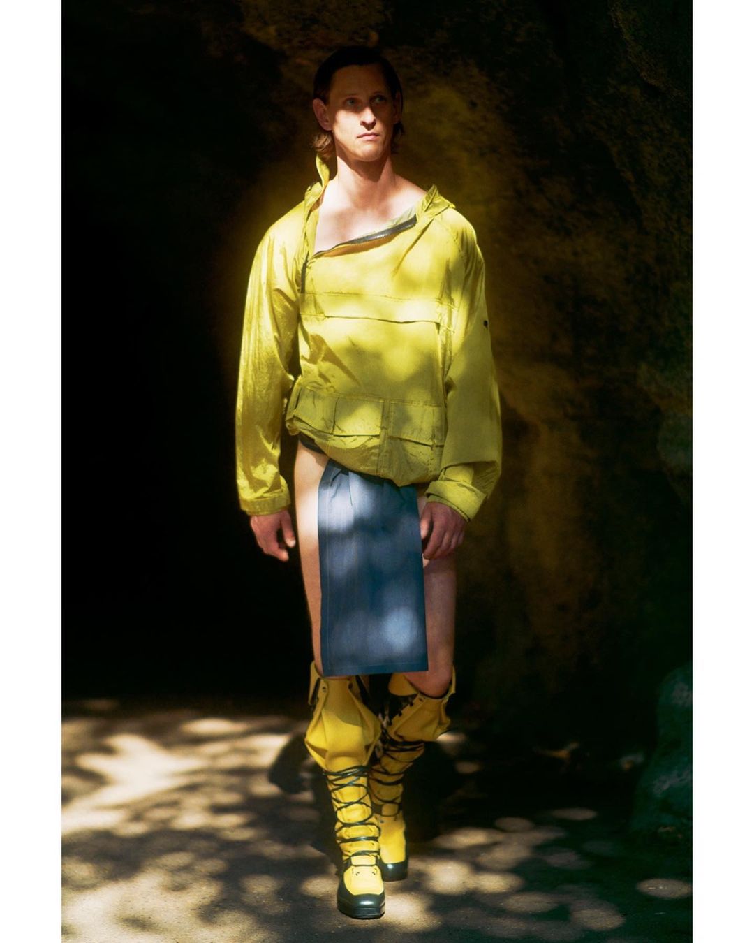 Male fashion model wears yellow hooded jacket, blue drape, and yellow boots in a dimly lit cave. Fashion shot features unique attire.