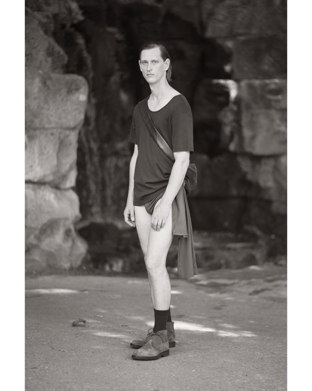 Monochrome full shot of a slender adult man in a dark t-shirt and shoes standing outdoors in front of a stone wall looking at the camera.