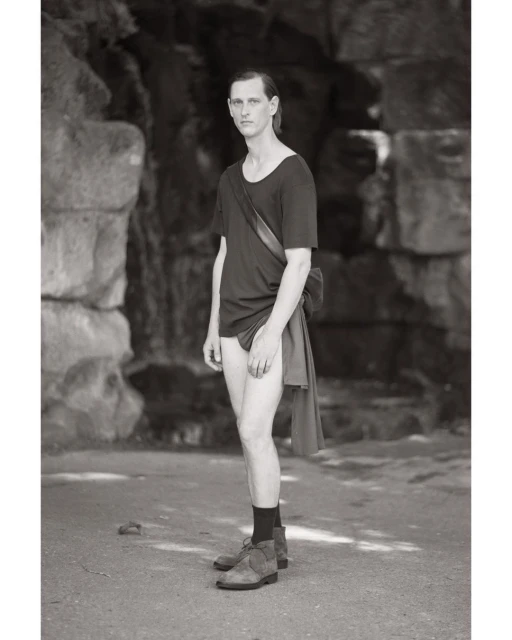 Monochrome full shot of a slender adult man in a dark t-shirt and shoes standing outdoors in front of a stone wall looking at the camera.