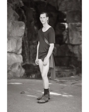 Monochrome full shot of a slender adult man in a dark t-shirt and shoes standing outdoors in front of a stone wall looking at the camera.