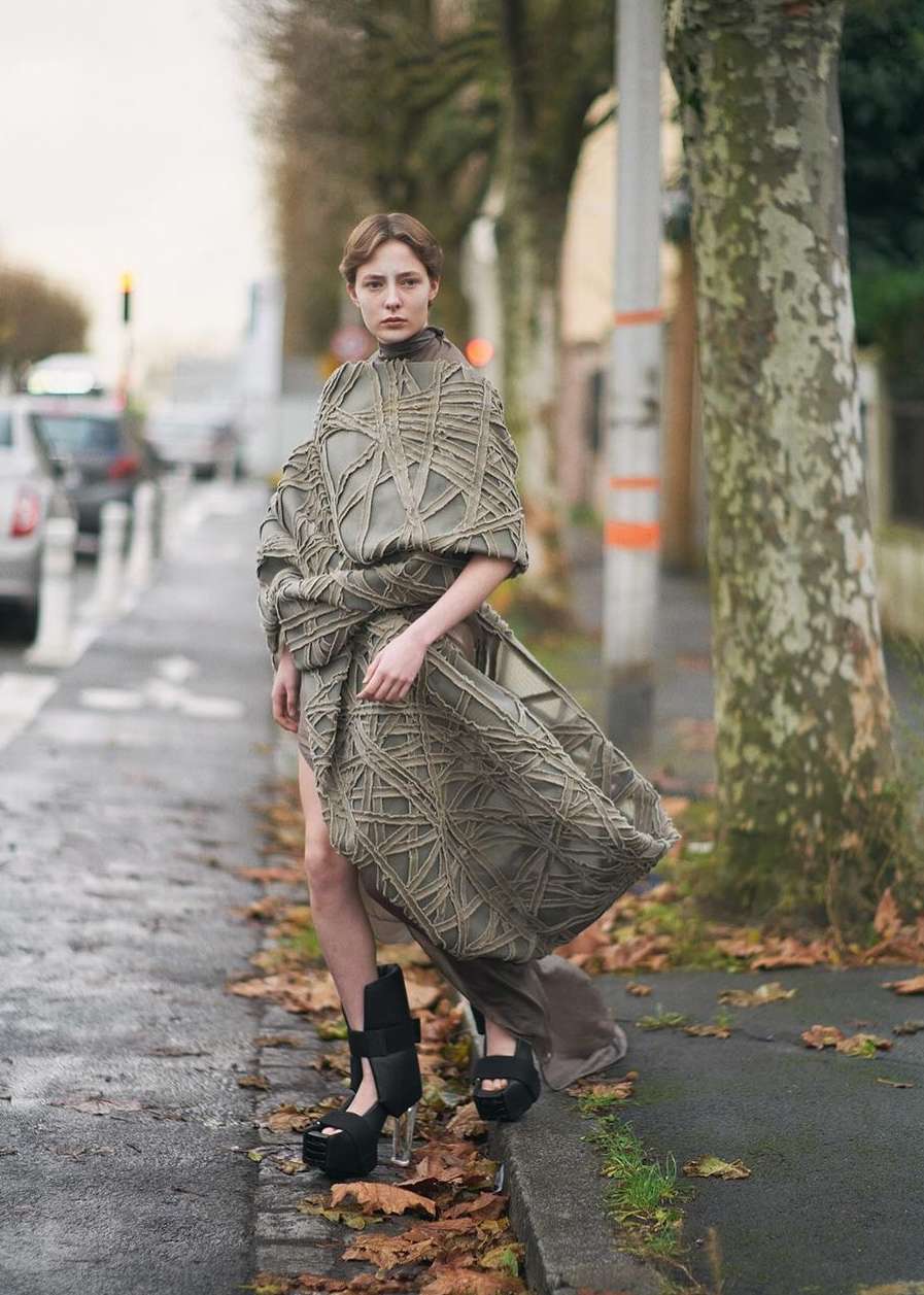 Woman wearing a conceptual gray woven dress and platform heels stands on a city street with bare trees in a fashion editorial photo.