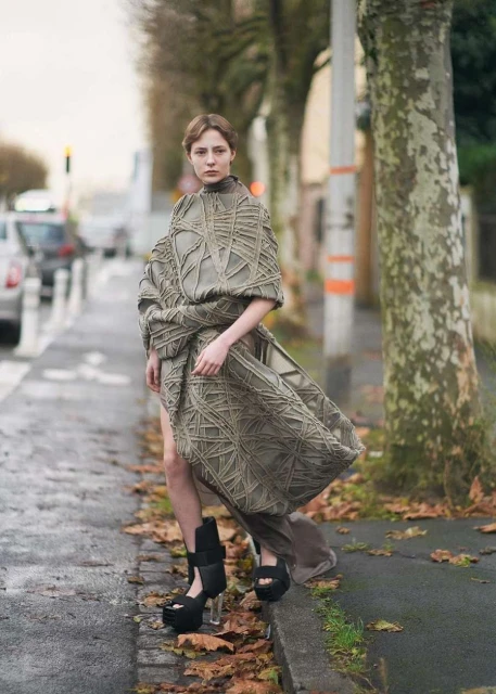 Woman wearing a conceptual gray woven dress and platform heels stands on a city street with bare trees in a fashion editorial photo.