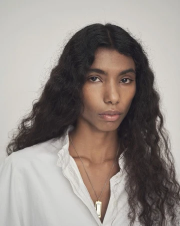 Portrait of a woman with long dark wavy hair wearing a white collared shirt and a pendant necklace against a light grey background.