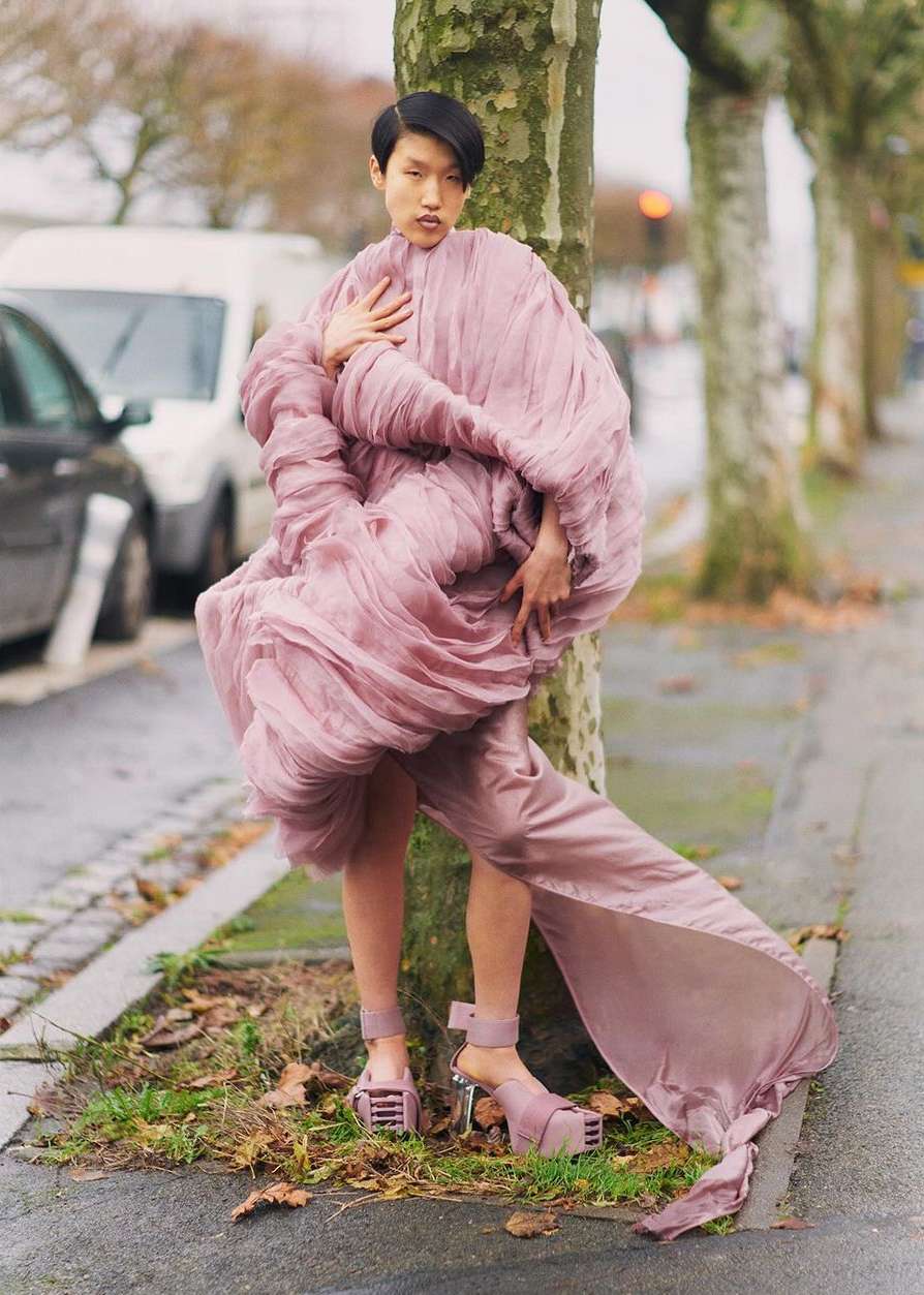 A stylish Asian woman in a lilac dress posing by a tree on a city street with cars and buildings visible in the background on an overcast day.