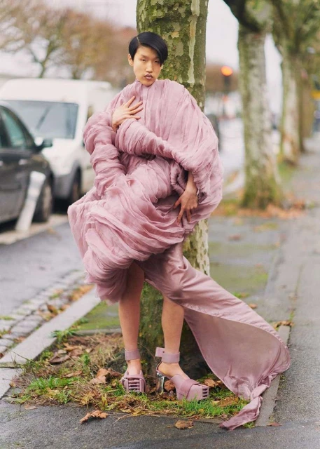 A stylish Asian woman in a lilac dress posing by a tree on a city street with cars and buildings visible in the background on an overcast day.