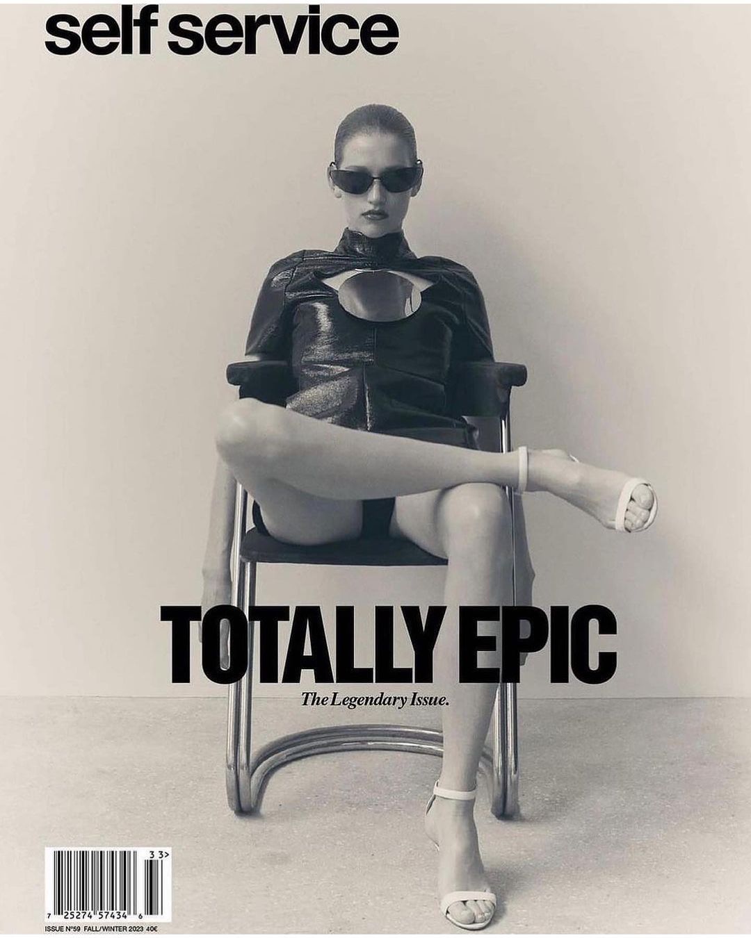 Black and white magazine cover shot of a woman in sunglasses and an avant-garde black outfit, sitting on a tubular metal chair.