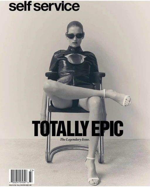 Black and white magazine cover shot of a woman in sunglasses and an avant-garde black outfit, sitting on a tubular metal chair.