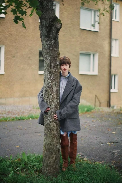 Adult posing behind a tree wearing a gray overcoat and brown, over-the-knee boots with an apartment building in the background.
