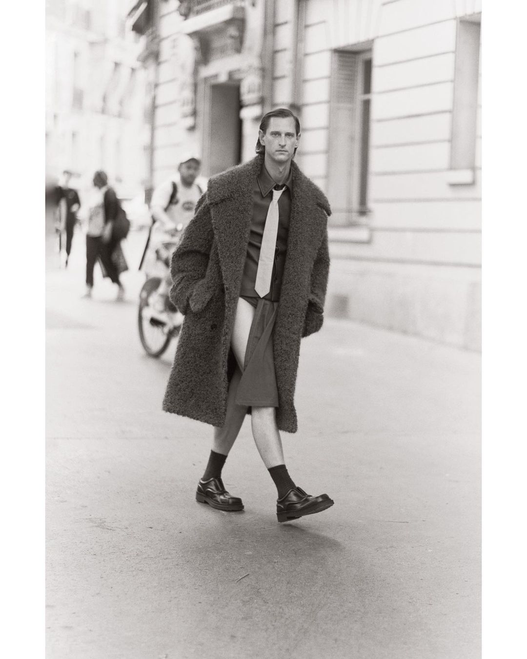 Monochrome street-style fashion shot: A male model in an oversized coat, shirt, tie, and shorts walks on a city street with a cyclist.